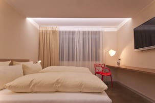 Una habitación de hotel con cama, una silla roja, escritorio, televisor y ventana con cortinas.