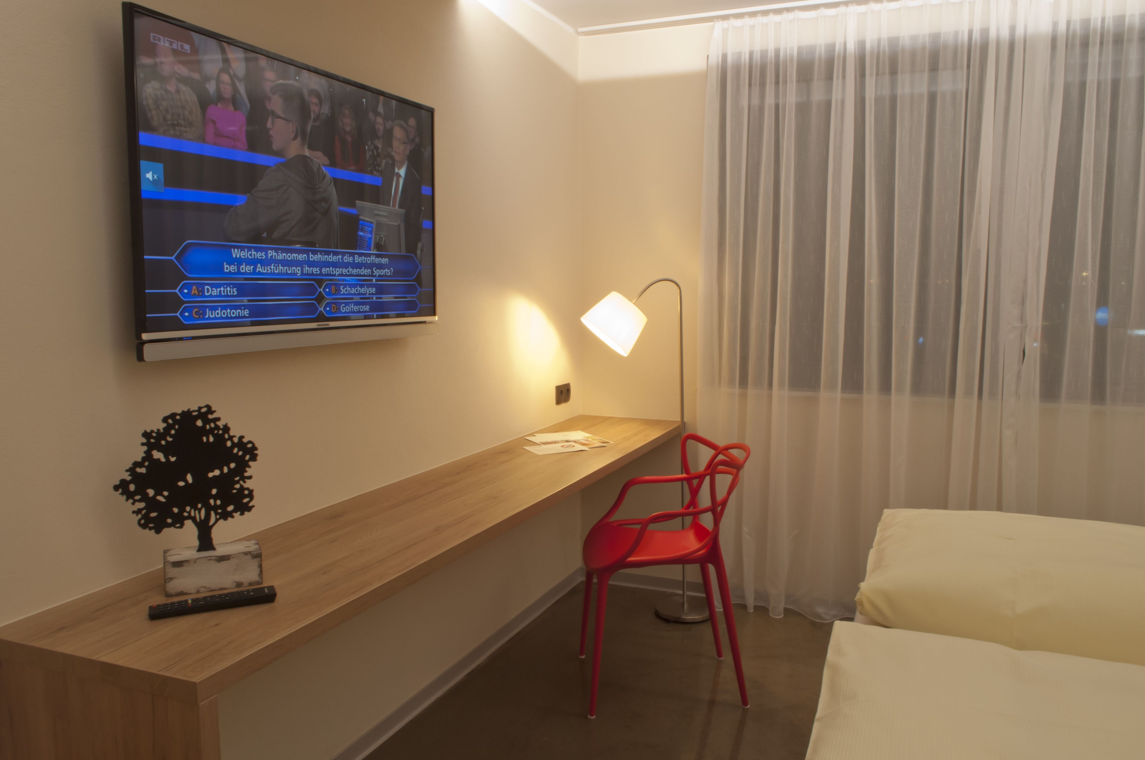 A hotel room with a wooden desk, a red chair, a TV mounted on the wall, and a bed with white bedding.