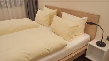 A neatly made bed with a wooden headboard, a bedside table with a lamp, and a window with sheer curtains.