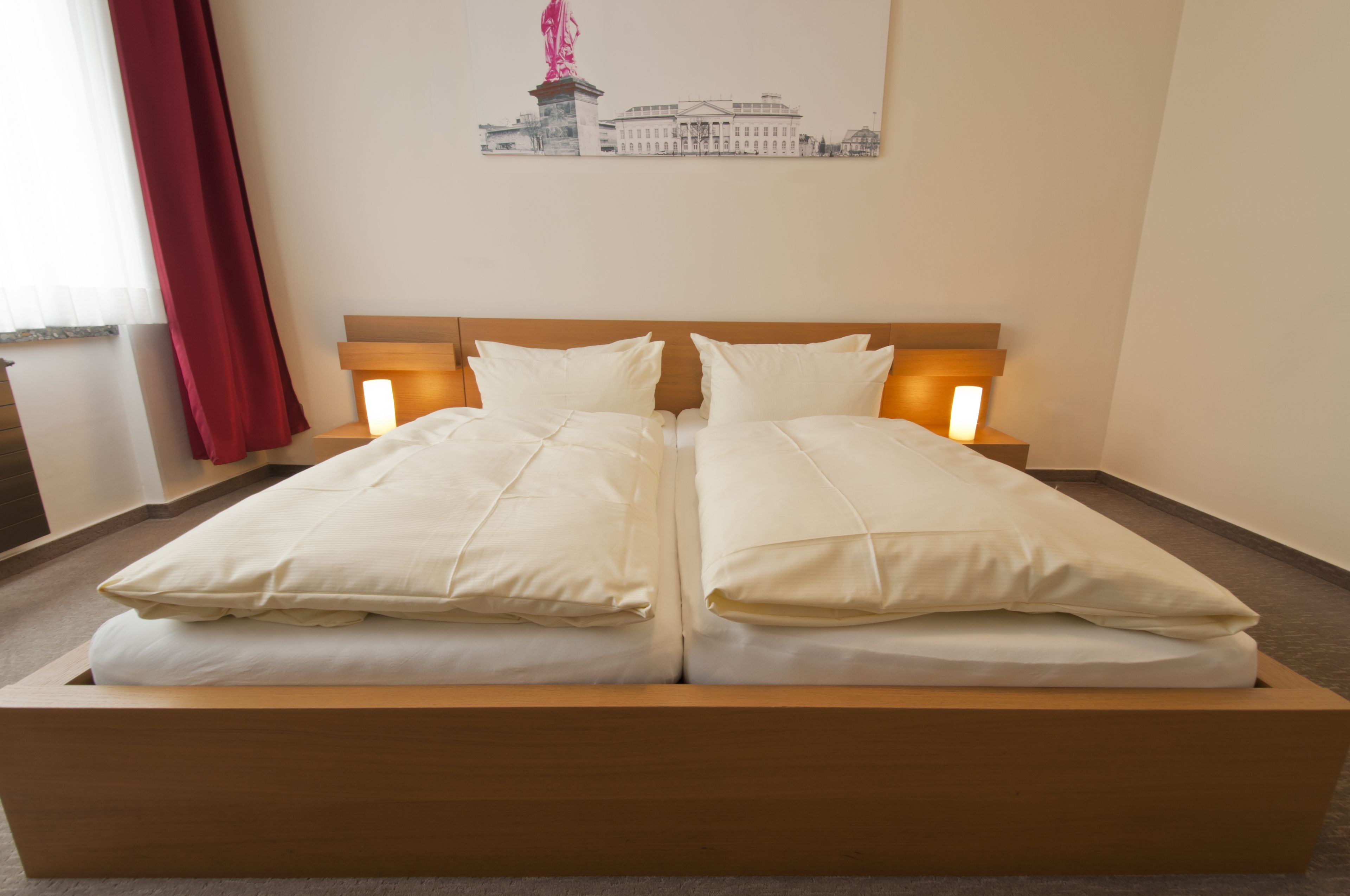 A double bed with white bedding and wooden headboard, a red curtain, and a wall-mounted artwork of a building.