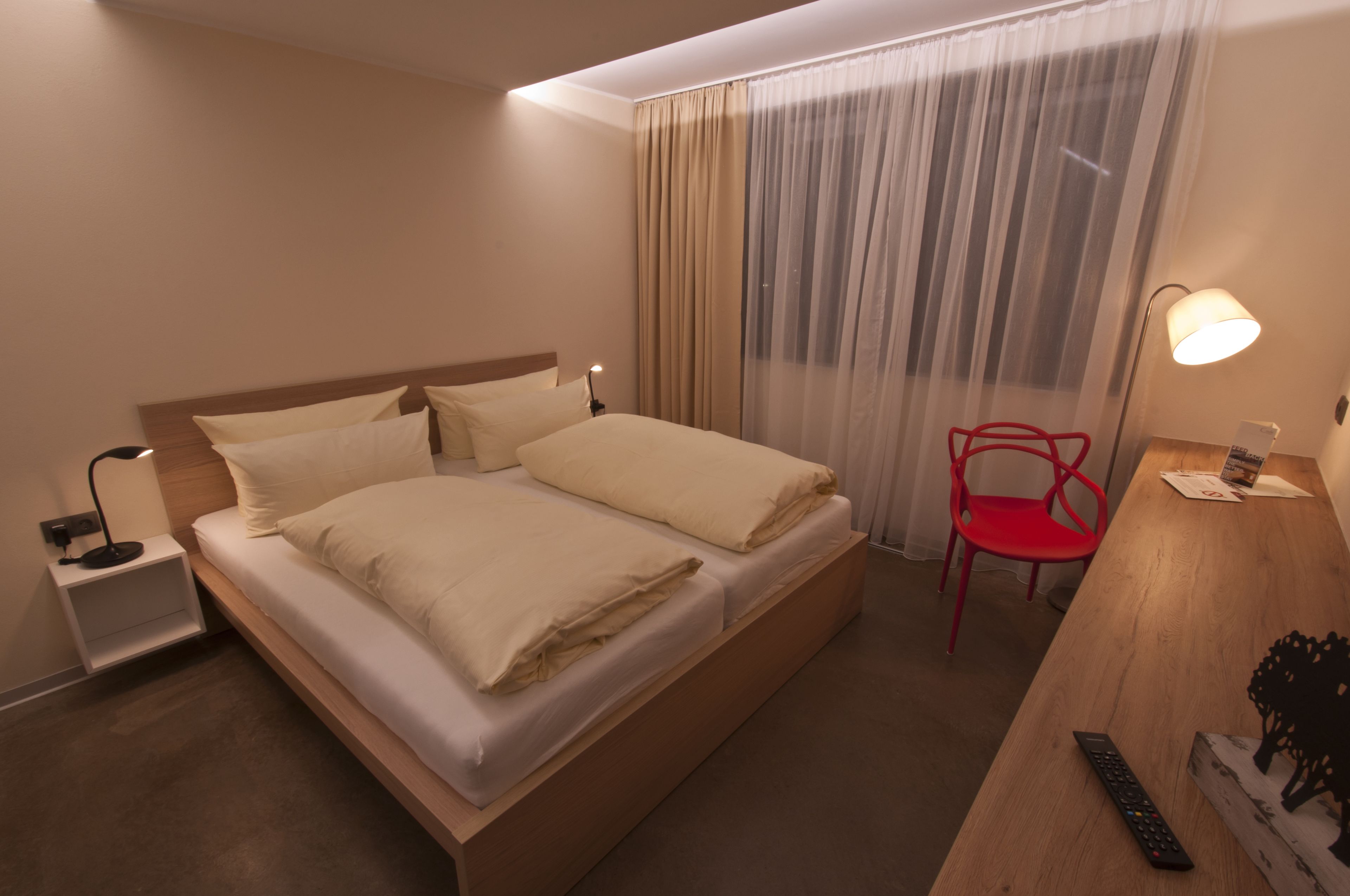 A hotel room with a bed, bedside table, lamp, chair, and a desk with a remote control.
