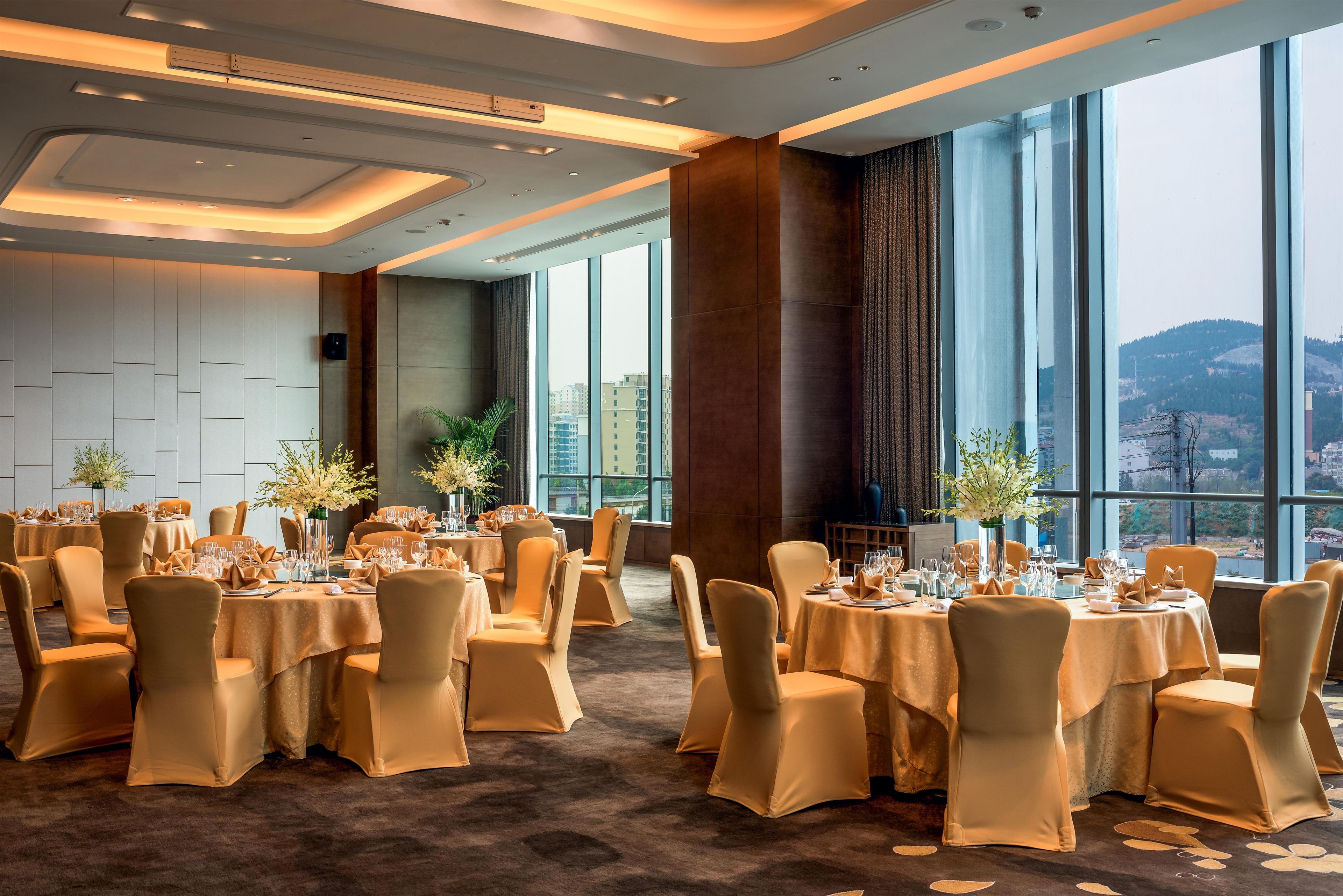 Un amplio salón de banquetes con mesas redondas dispuestas para cenar, grandes ventanales con vistas a la ciudad y un diseño de techo moderno.