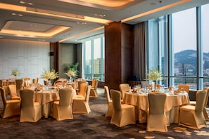 Un amplio salón de banquetes con mesas redondas dispuestas para cenar, grandes ventanales con vistas a la ciudad y un diseño de techo moderno.