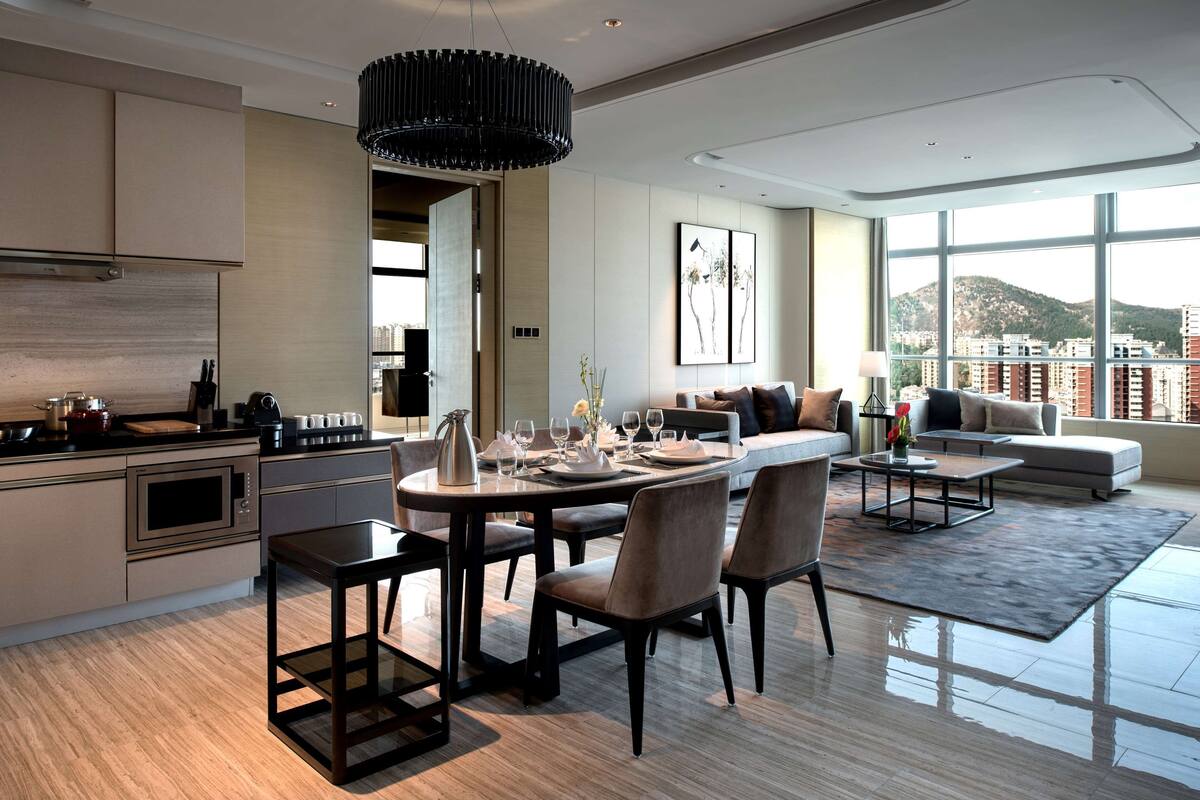 A modern kitchen with a dining area, a living room with a sofa and a coffee table, and large windows offering a city view.