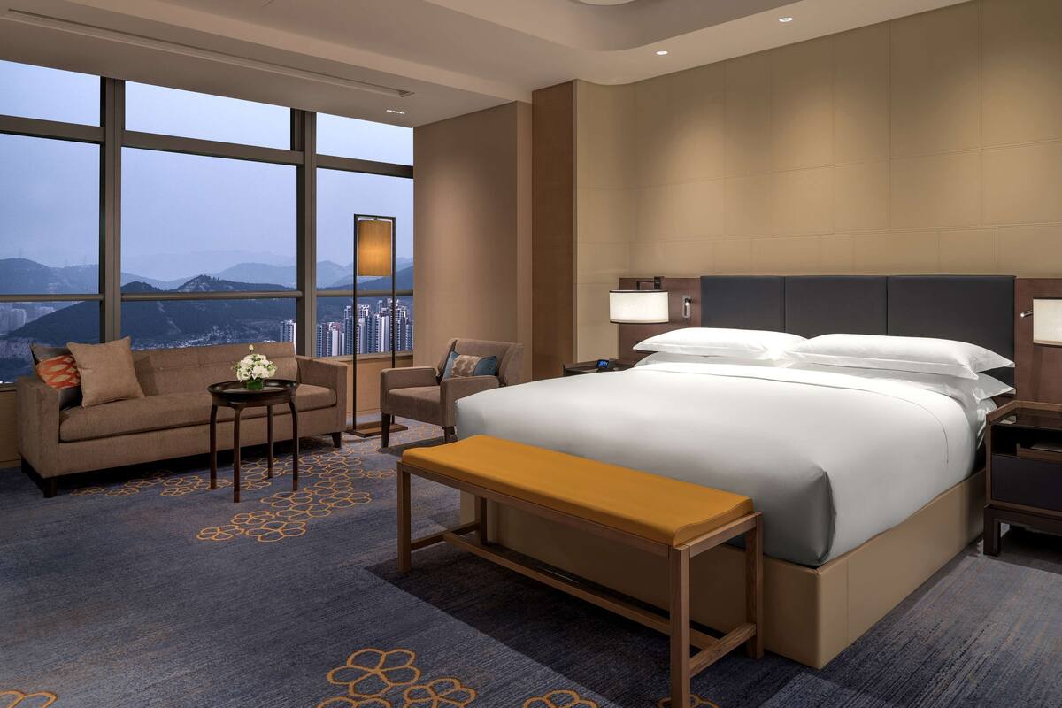 A hotel room with a large bed, a sofa, a small table, and a bench. The room has large windows offering a view of the city and mountains.