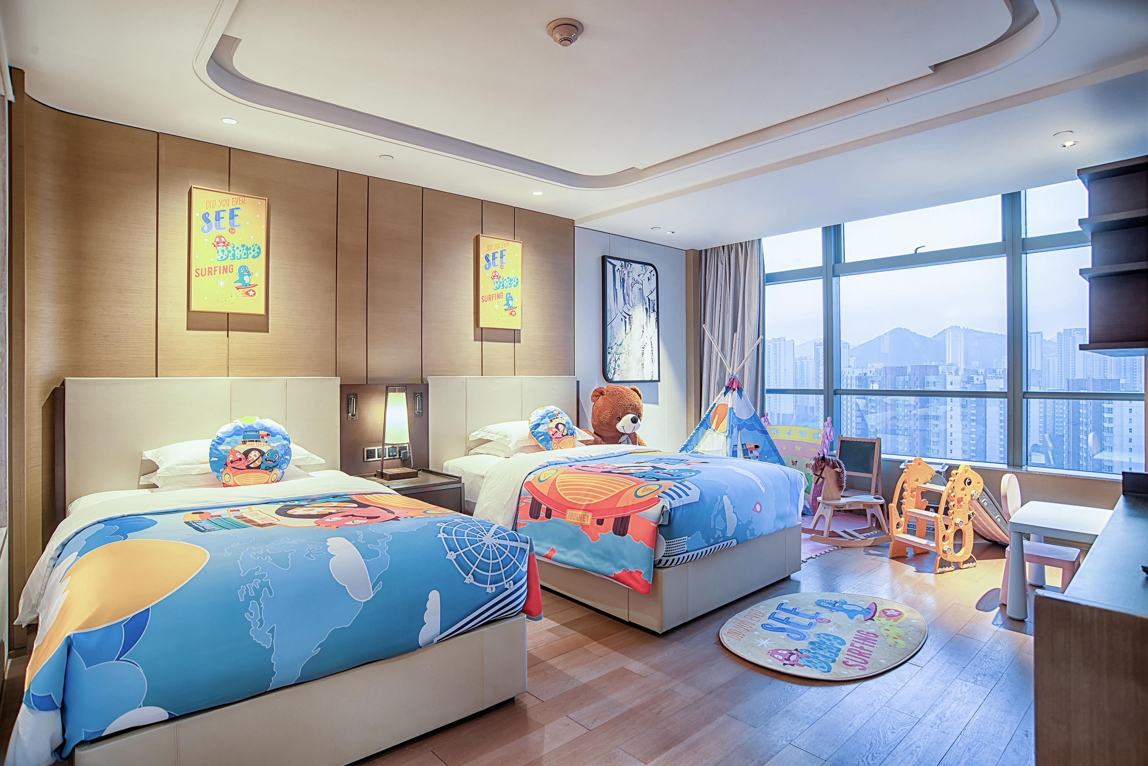 A hotel room with two beds, a desk, a chair, a window with a city view, and a teddy bear.
