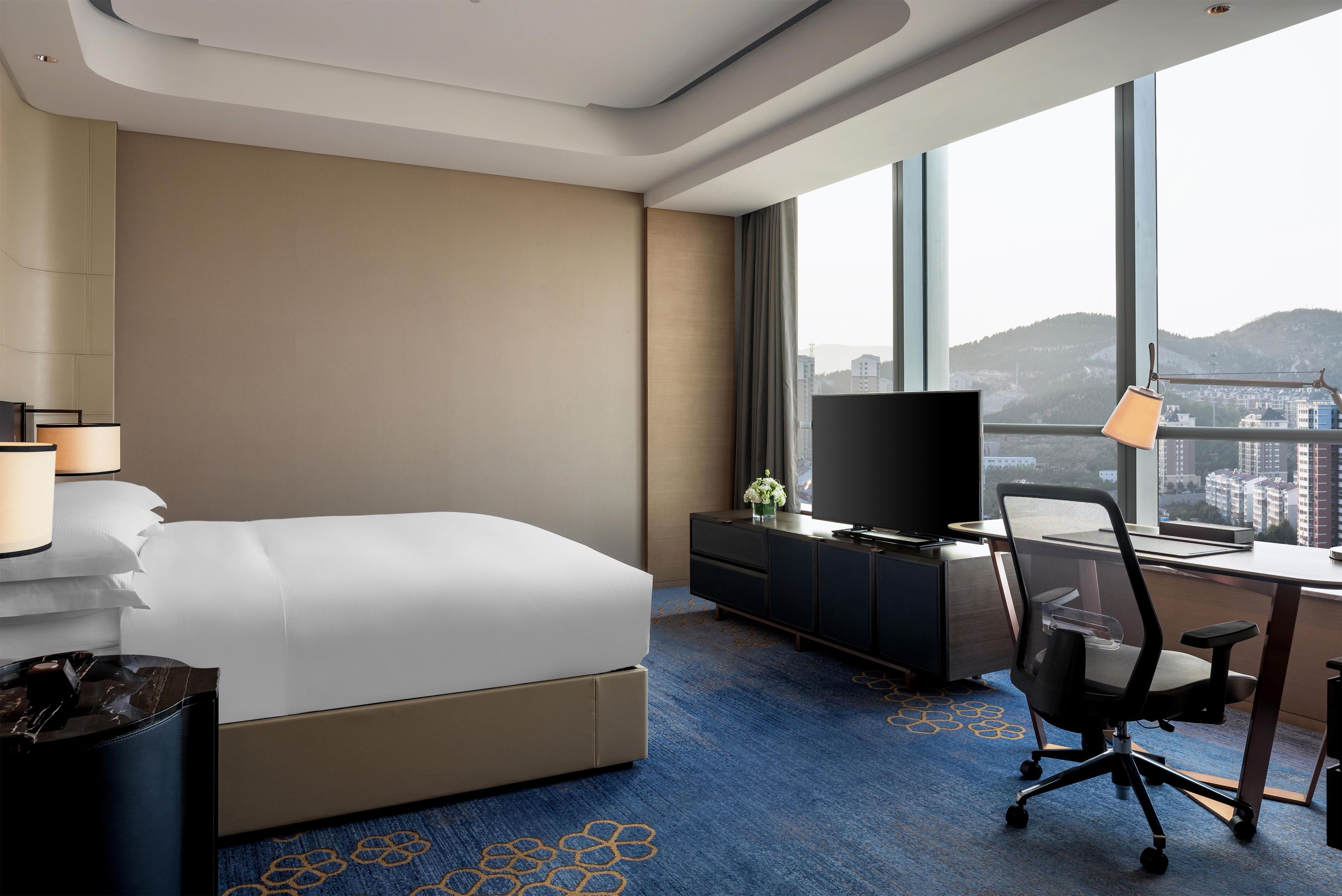 Habitación de hotel con cama, escritorio con silla, televisor y un amplio ventanal con vista a la ciudad.