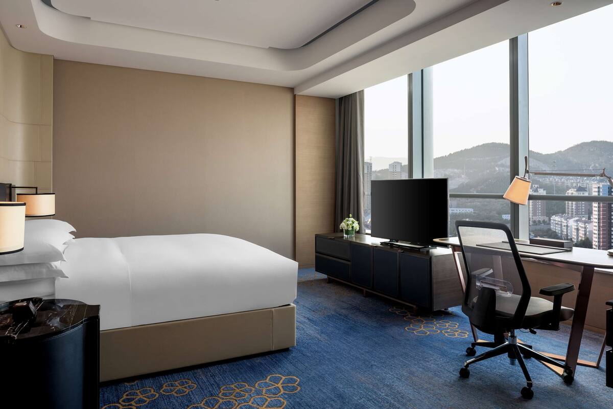 A hotel room with a bed, a desk with a computer, a TV, and a large window with a city view.