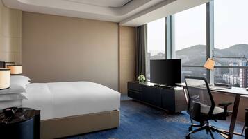 A hotel room with a bed, a desk with a computer, a TV, and a large window with a city view.