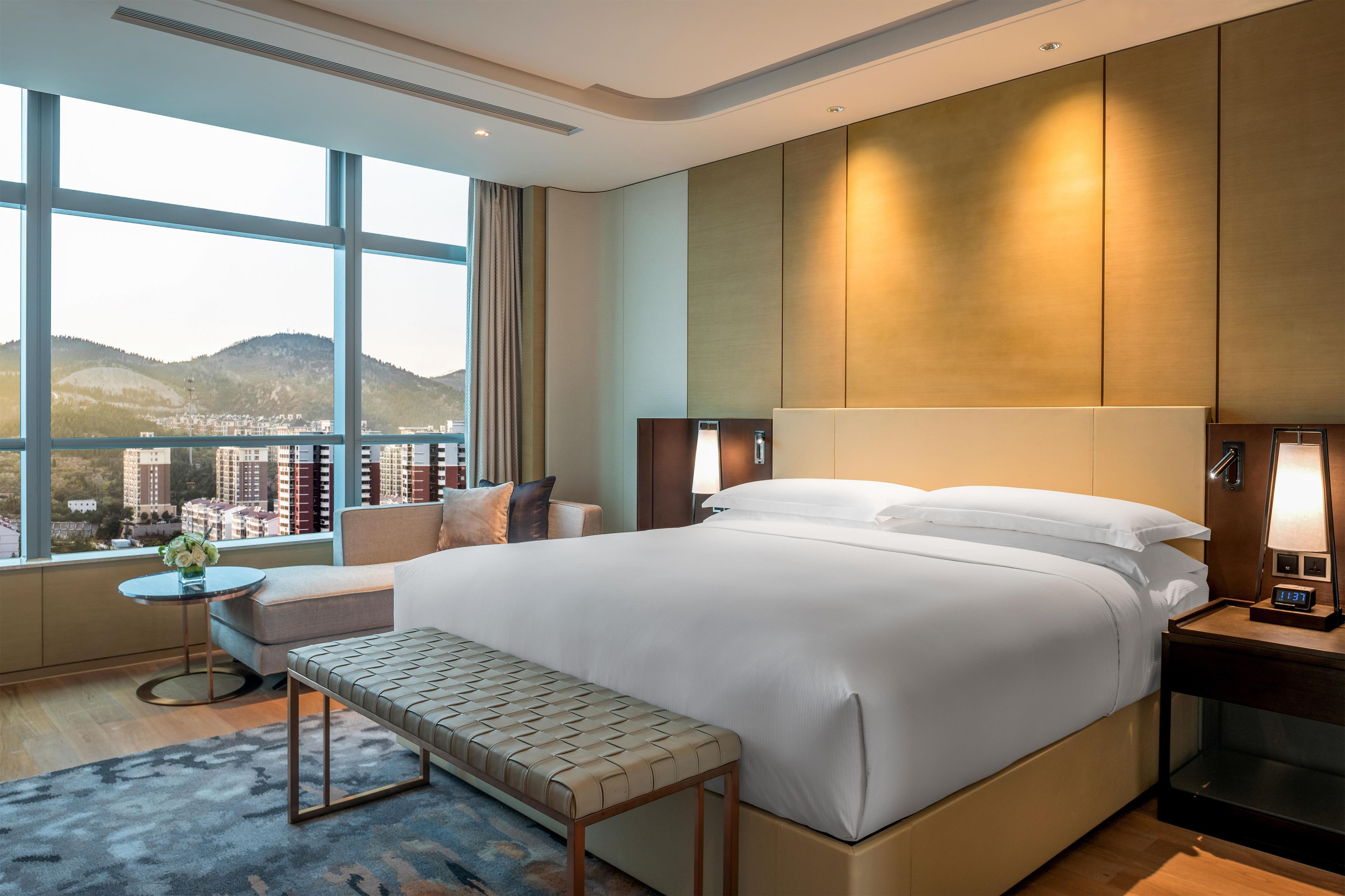 Quarto de hotel com cama grande, mesa de cabeceira, um banco, vista da cidade e mesa de apoio com abajur.