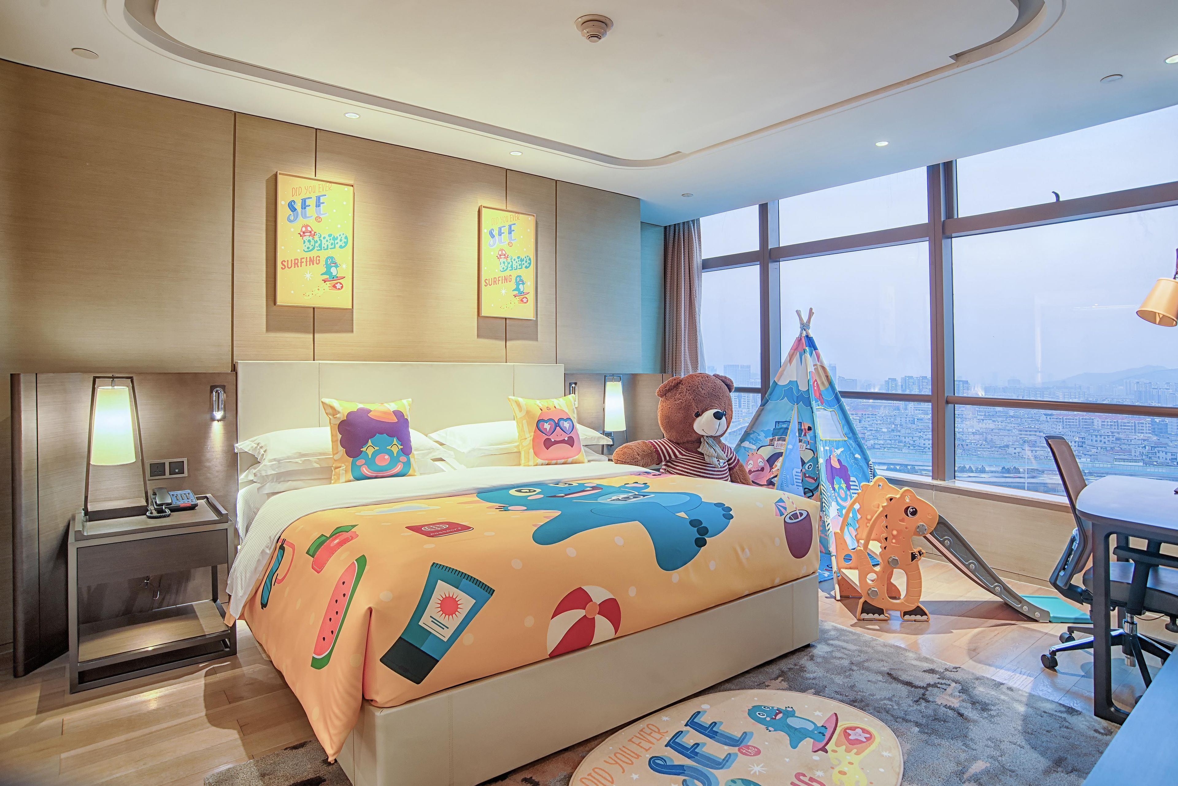 Una habitación de hotel con una cama, un escritorio, una silla, un osito de peluche, una tienda de campaña y vistas de la ciudad.
