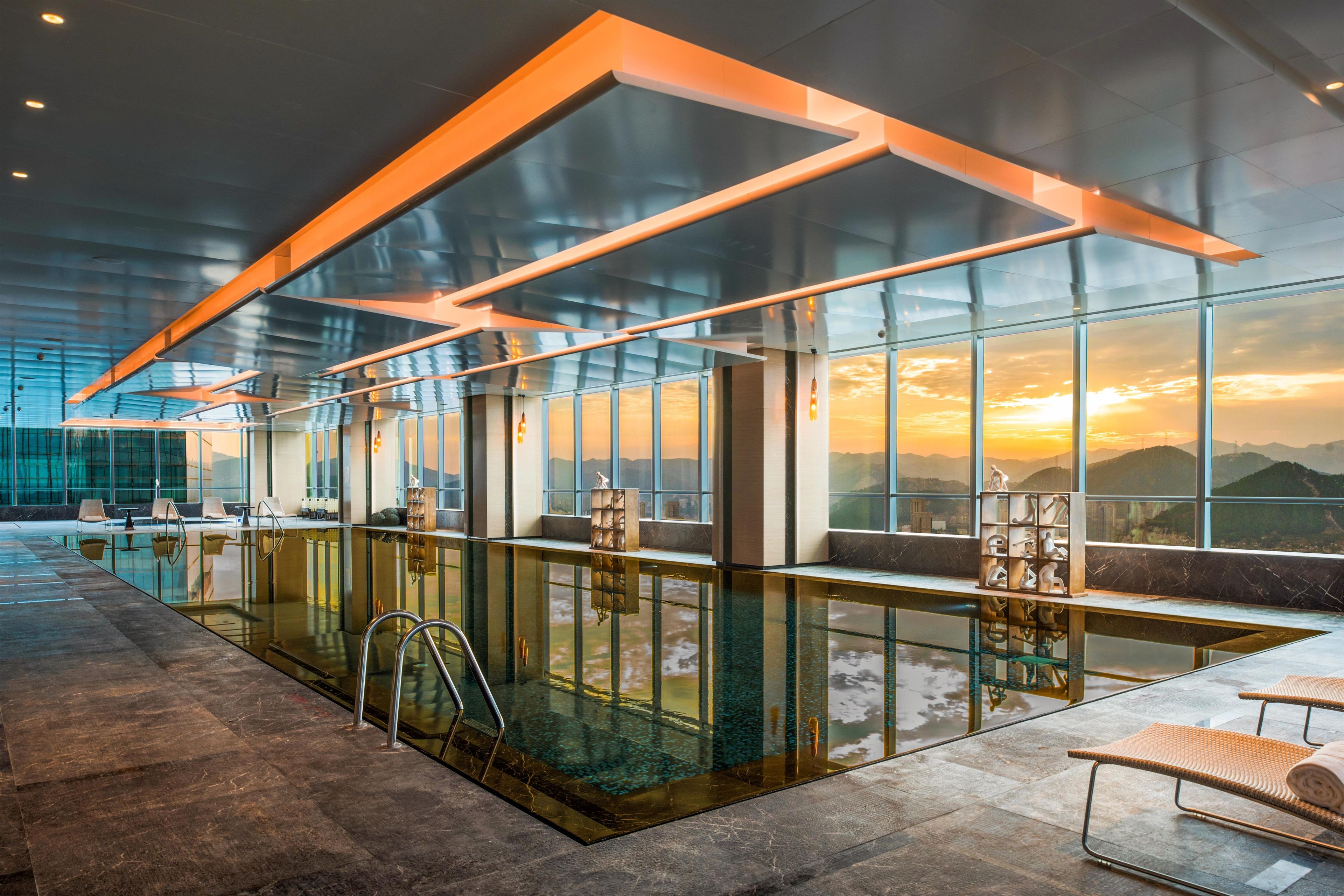 A modern indoor pool area with glass walls, a clear view of the sunset, and a seating area with lounge chairs.