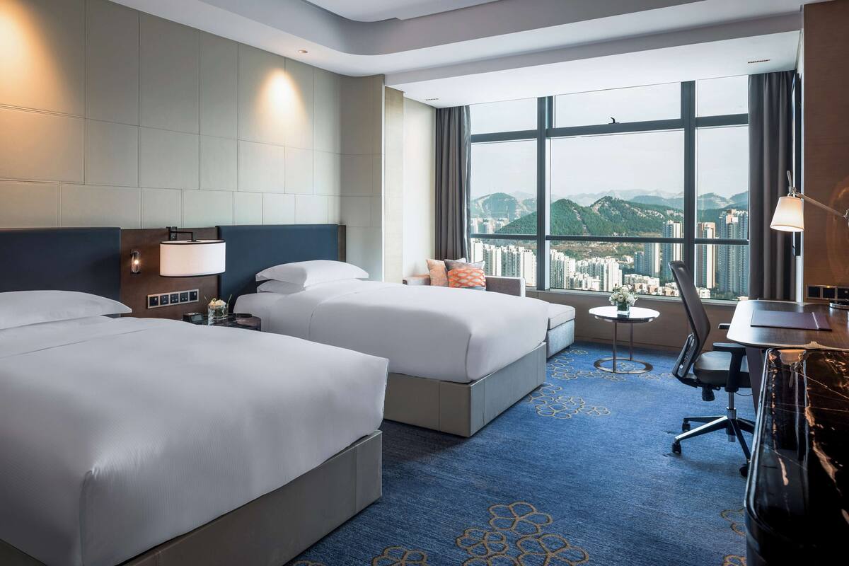 A hotel room with two beds, a desk, a chair, and a large window with a city view.