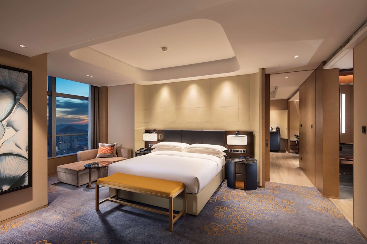 A modern hotel room with a large bed, bedside tables, a bench, a view of the cityscape, and a large abstract painting.