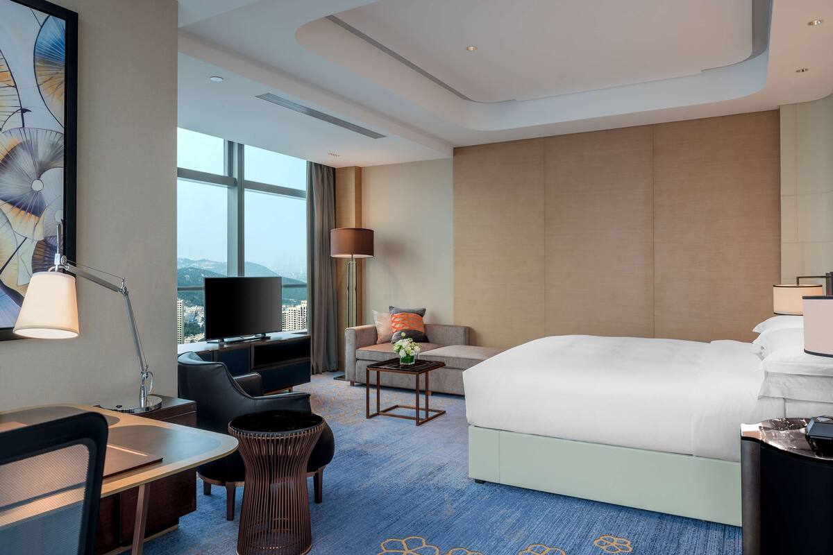 A hotel room with a bed, a desk, a chair, a TV, and a view of the city.