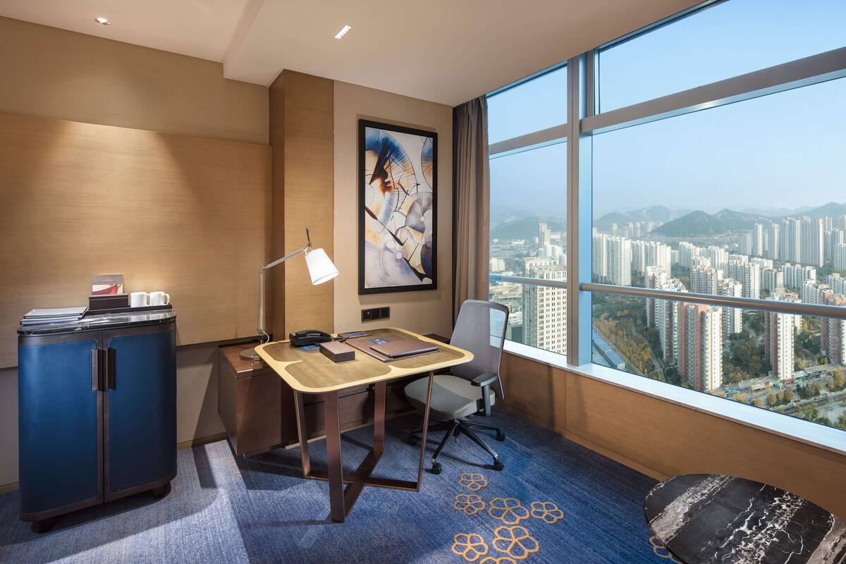 A modern hotel room with a large window offering a city view, a desk with office supplies, a blue cabinet, and a decorative rug.