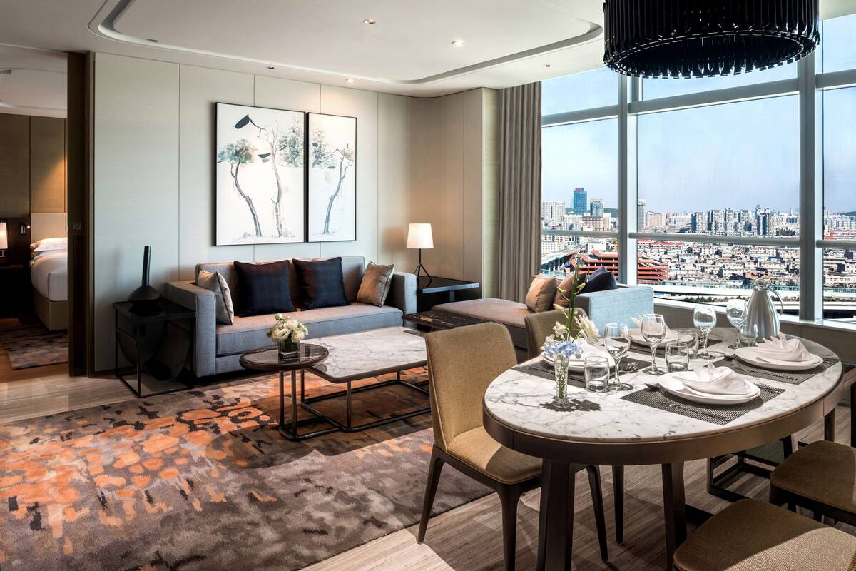 A modern hotel room with a city view, a sofa, a dining table set for two, and a large window.