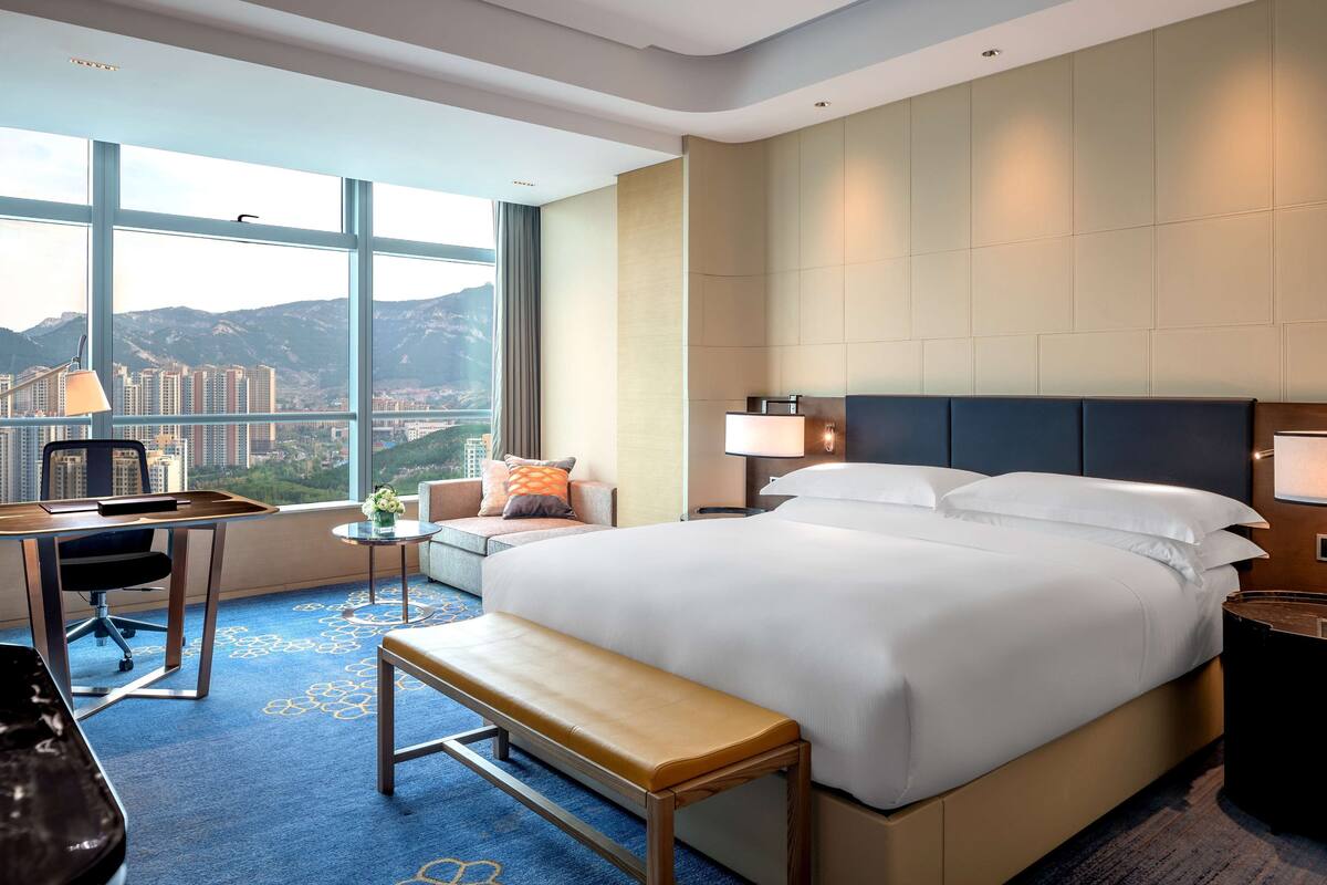 A hotel room with a large bed, a desk, a bench, and a view of a cityscape.