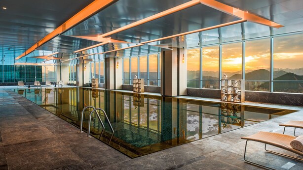 A modern indoor pool area with glass walls, a lounge with seating, and a view of the sunset through large windows. - Hilton Jinan South Hotel & Residences (Jinan)