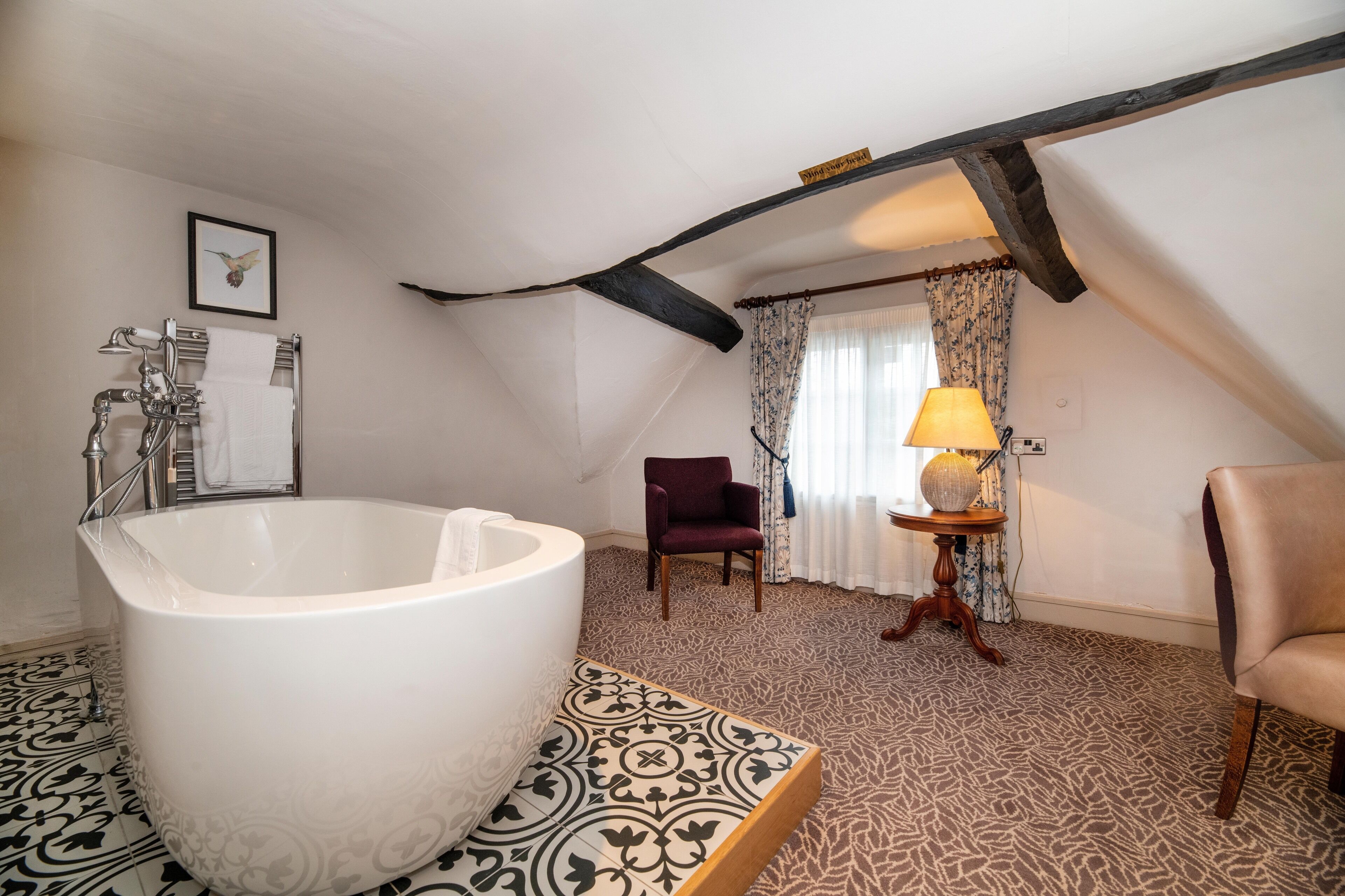 A bathroom with a freestanding bathtub, a wooden chair, a small table with a lamp, and patterned floor tiles.