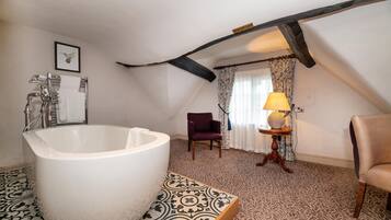 A bathroom with a freestanding bathtub, a wooden chair, a small table with a lamp, and patterned floor tiles.