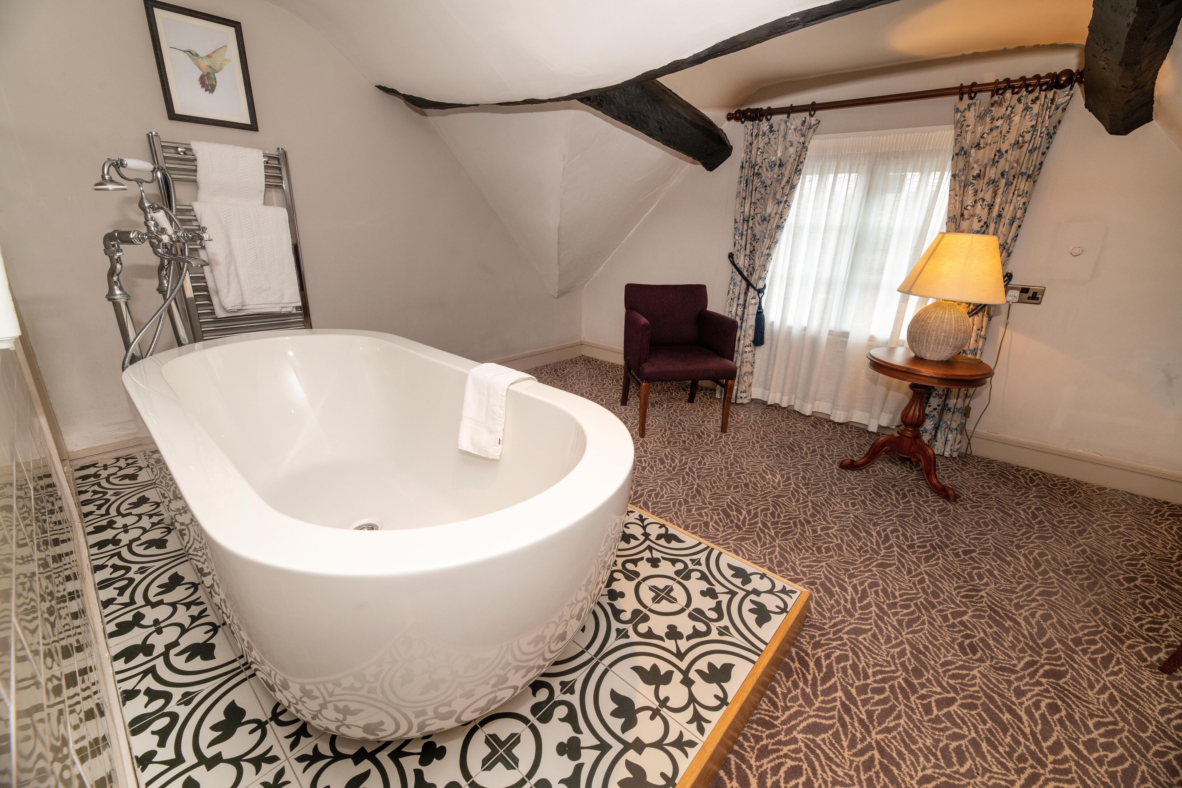 Ein Badezimmer mit freistehender Badewanne, einem Sessel, einem kleinen Tisch mit Lampe und fugenlosen Bodenfliesen.
