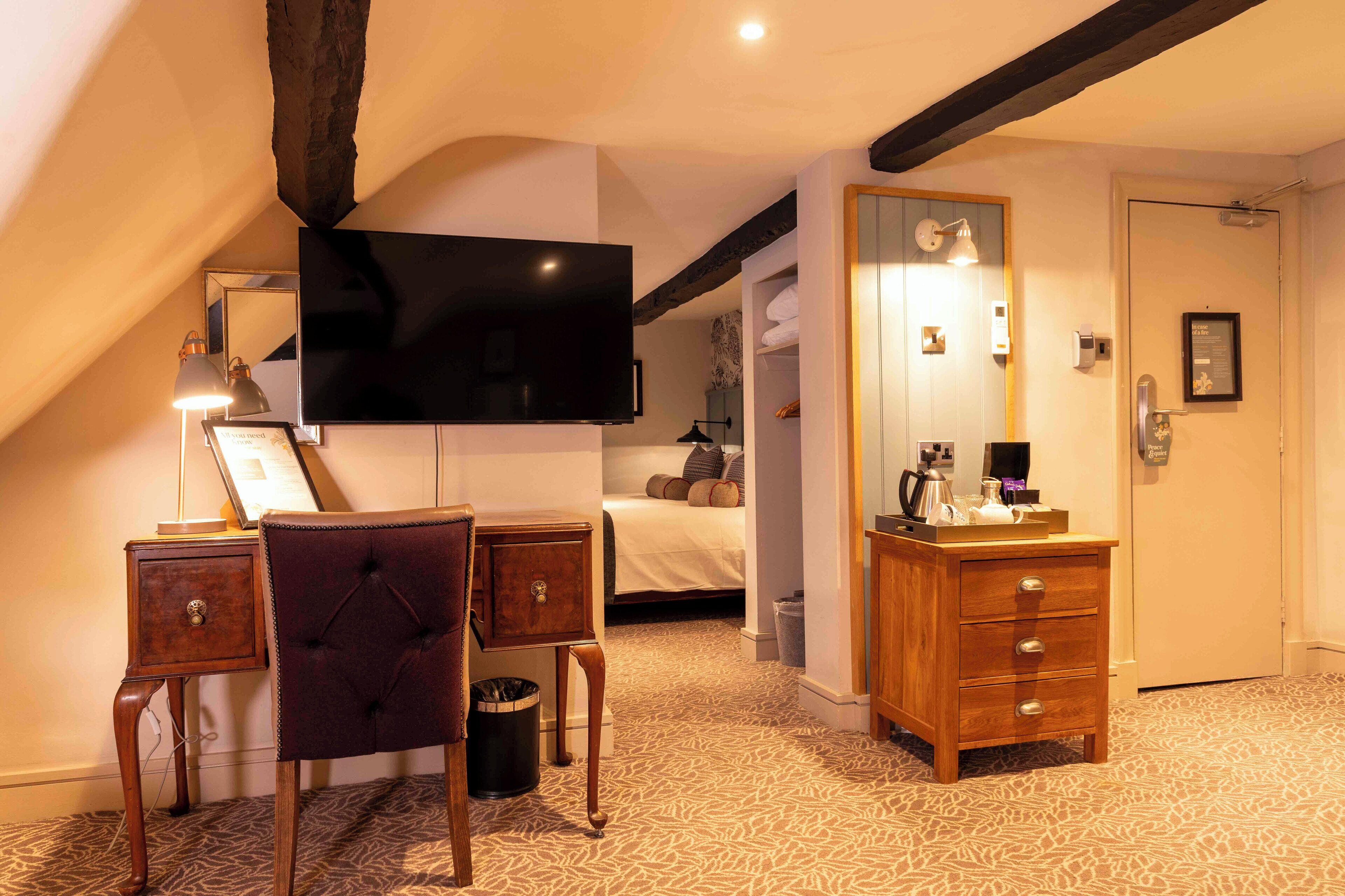 Habitación de hotel con un televisor de pantalla plana grande, un aparador de madera, una silla, un escritorio con lámpara, alfombra estampada y una cama con almohadas.
