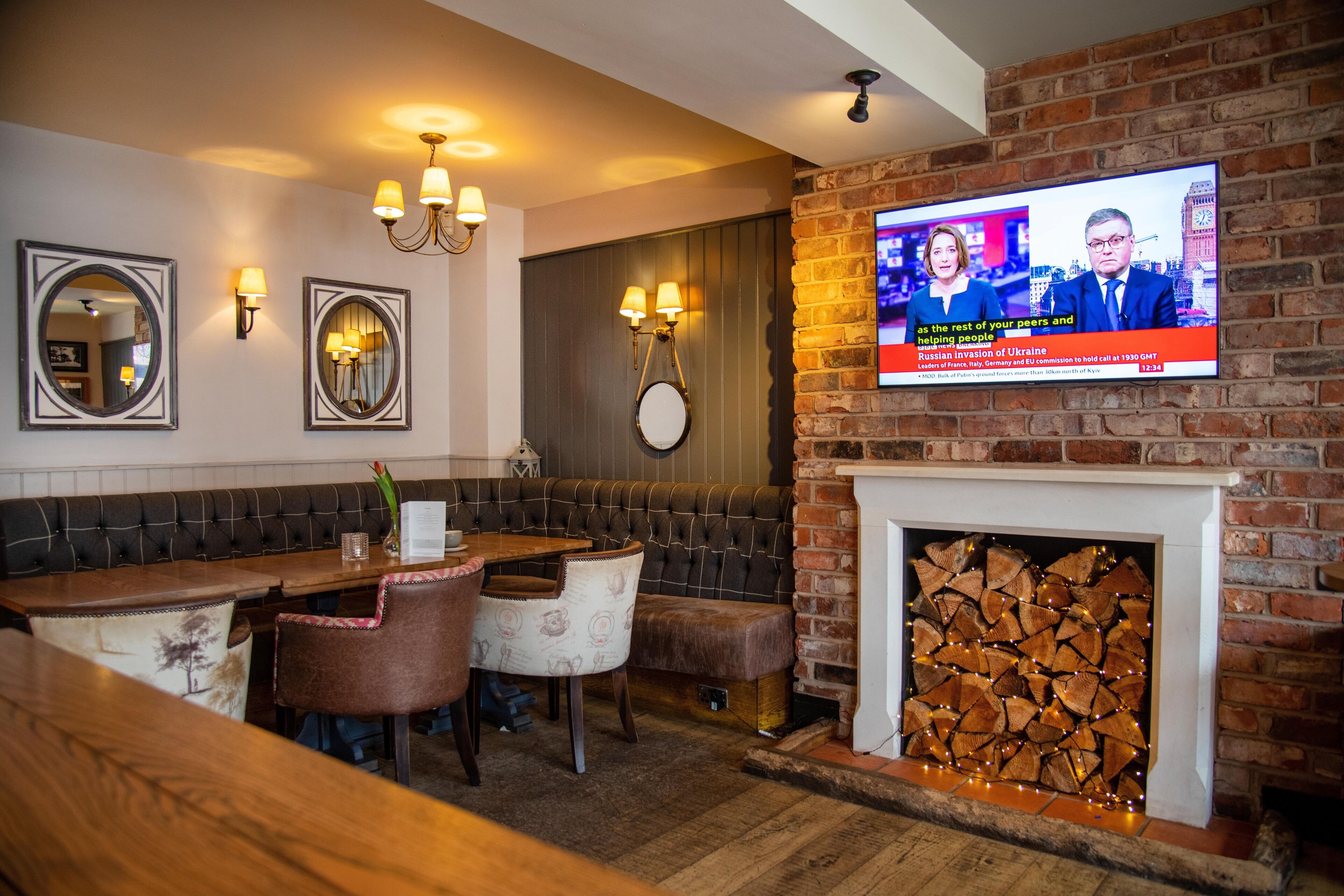 A cozy restaurant with a brick fireplace, a television displaying news, and a seating area with a wooden table and cushioned chairs.