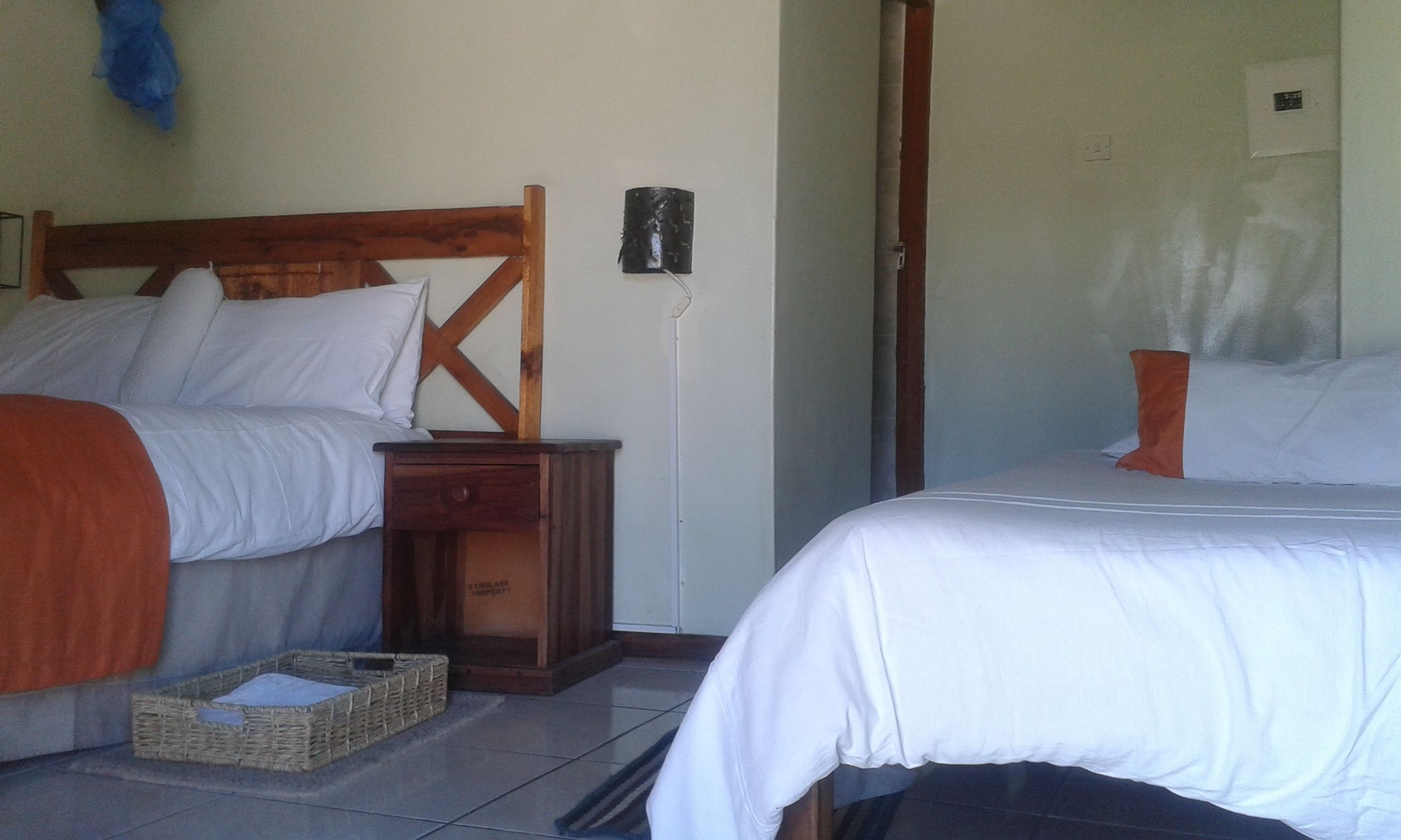 Una habitación con dos camas, cabecero de madera, una mesita de noche, una cesta y un aplique de pared.