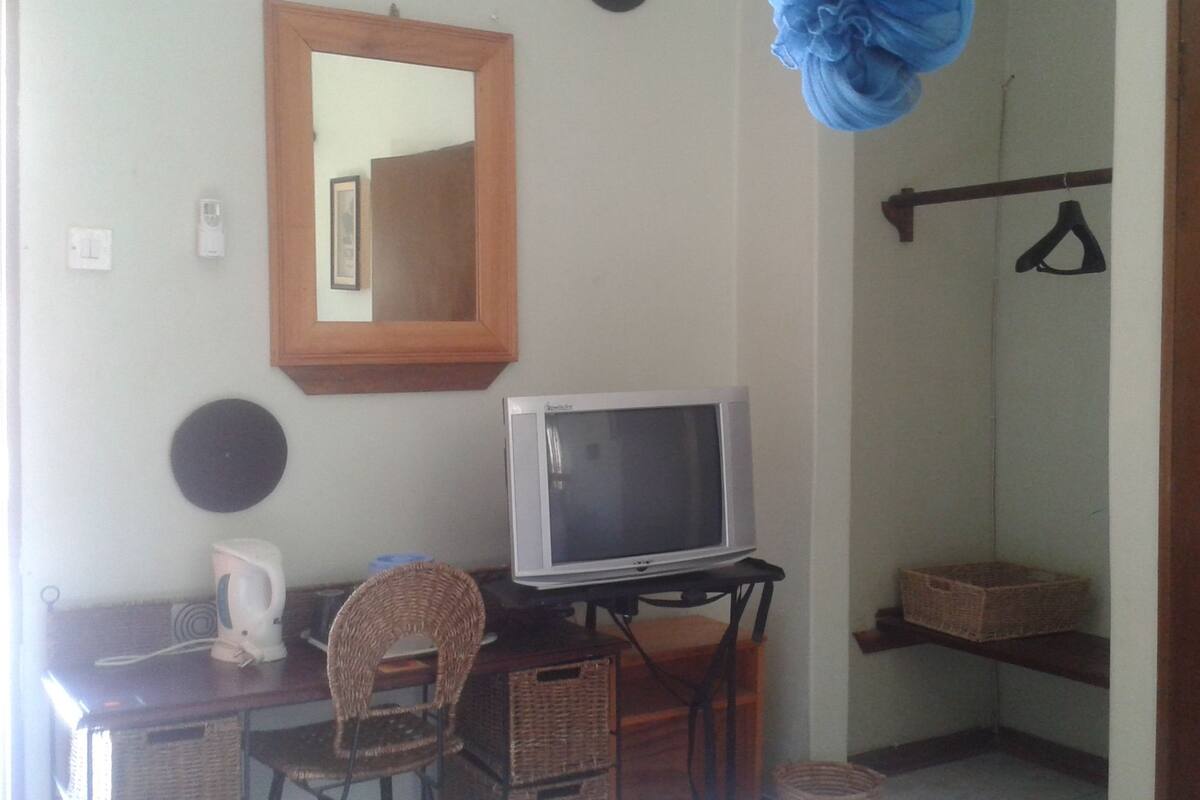 A small room with a television on a stand, a wicker chair, a mirror, and a shelf with baskets.