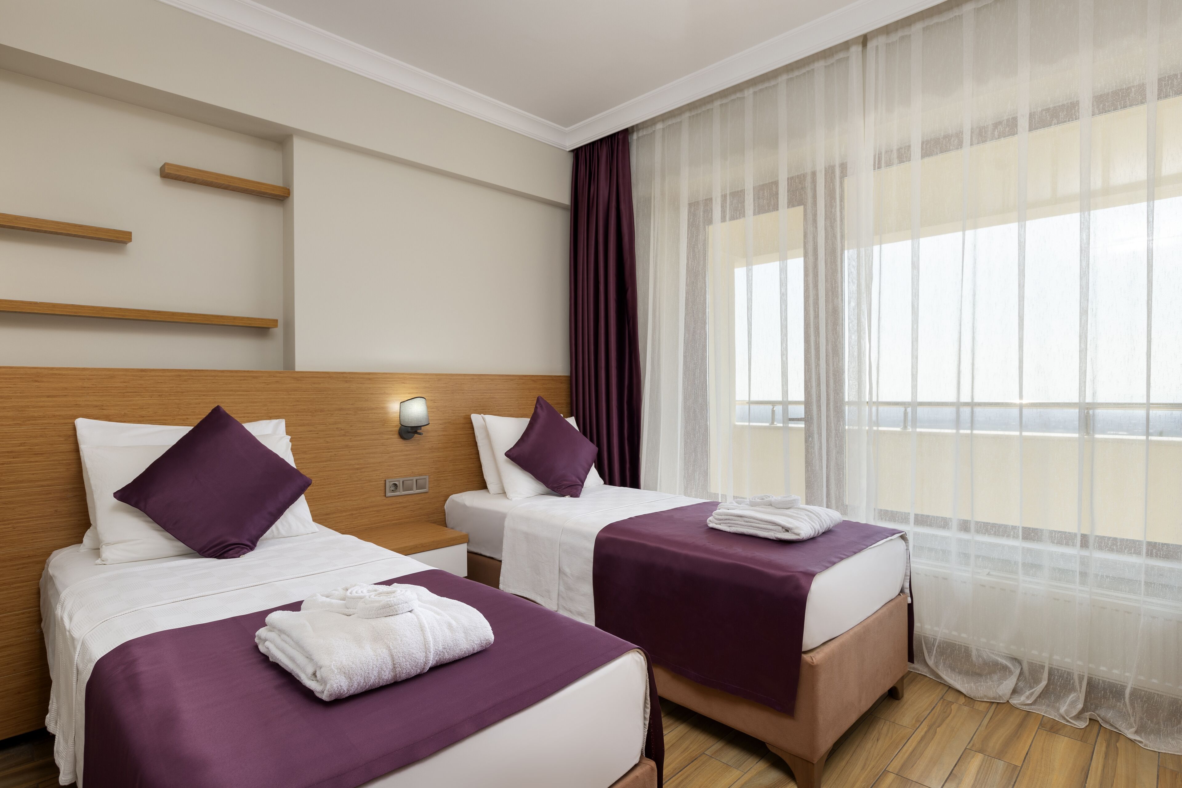 A hotel room with two beds, each with a purple bedspread, white pillows, and a white towel on the bed. There is a wooden headboard, a nightstand with a lamp, and a window with sheer curtains.