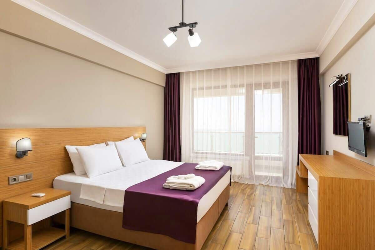 A hotel room with a wooden headboard, a bed with white linens, a purple throw blanket, a bedside table with a lamp, a wooden desk, and a TV on the wall.