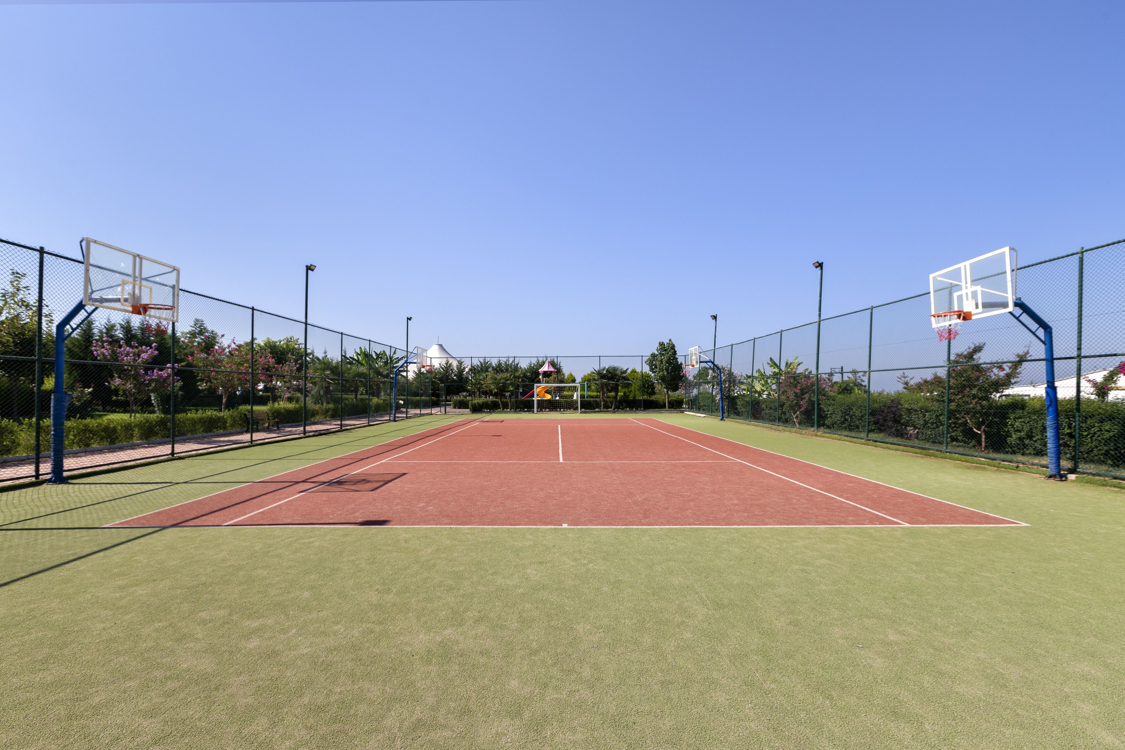 An outdoor tennis court with a net, surrounded by a fence, and a clear blue sky above.