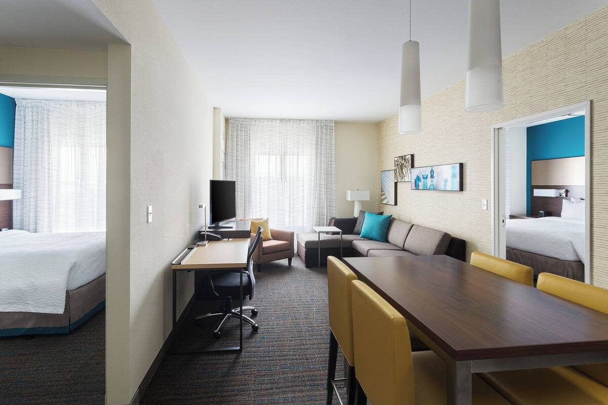 Una habitació d'hotel amb un llit, una taula de treball amb un ordinador, un sofà, una taula de dinar amb cadires i una finestra gran amb cortines.
