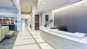A modern hotel reception area with a white reception desk, a seating area with a patterned rug, and a hallway leading to a restaurant. - Residence Inn by Marriott Houston West/Beltway 8 at Clay Rd. (Houston)