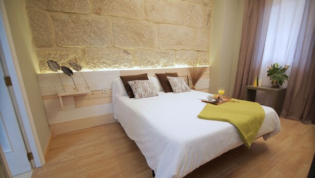 A bedroom with a stone wall, a bed with white bedding, patterned pillows, a bedside table with a lamp, and a small plant. - Alda Estación Vigo (Vigo)