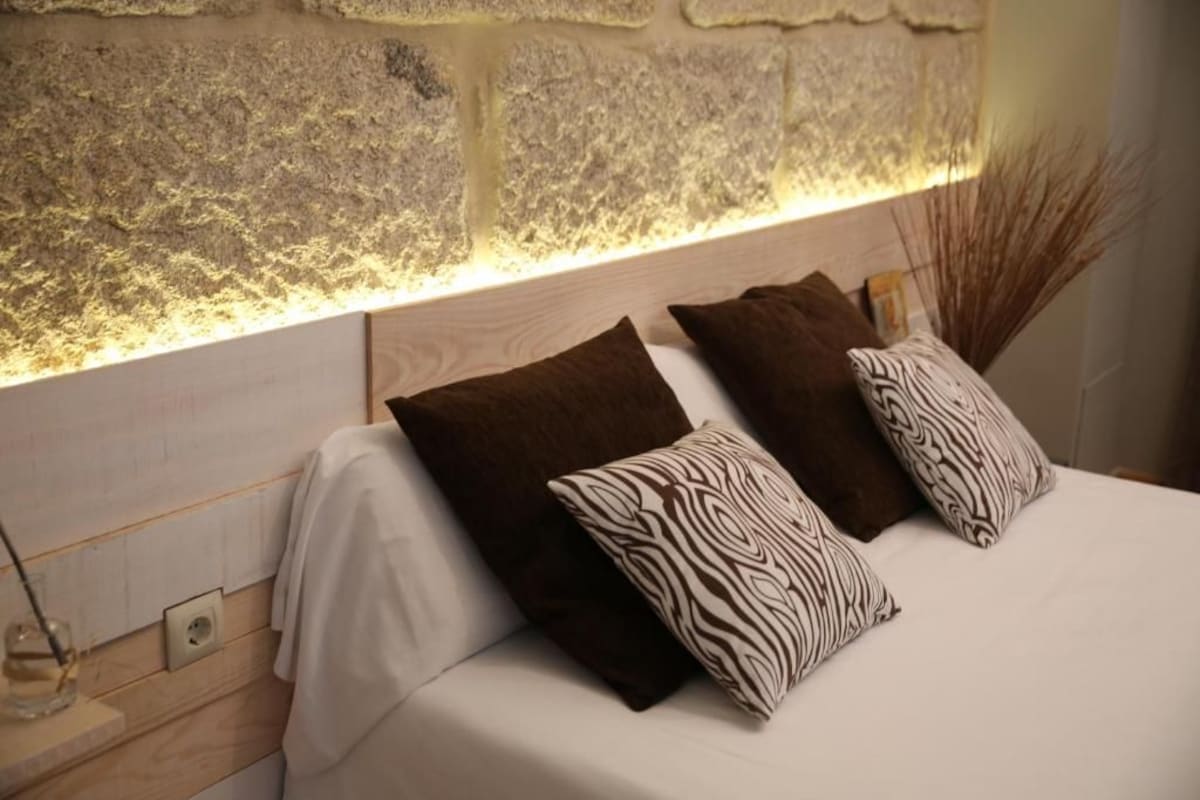 Una cama bien hecha con cabecera frente a una pared con textura, junto a una mesita de noche con vaso y vela.