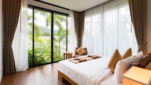 A hotel room with a large bed, a small table, and a view of greenery outside. - Nai Harn Baan-Bua Villas (Rawai)