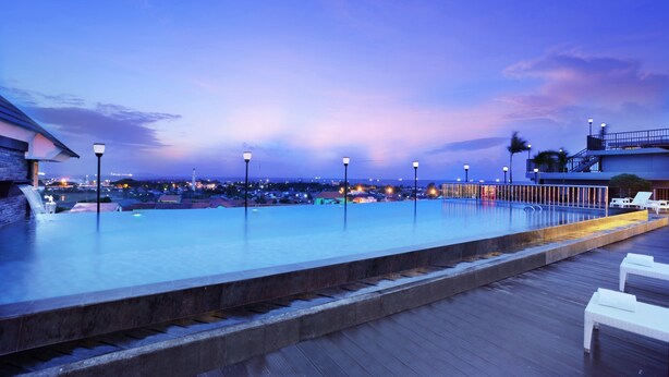 Una piscina infinita en la azotea con terraza de madera y sillones de descanso, con vistas a un paisaje urbano al anochecer.