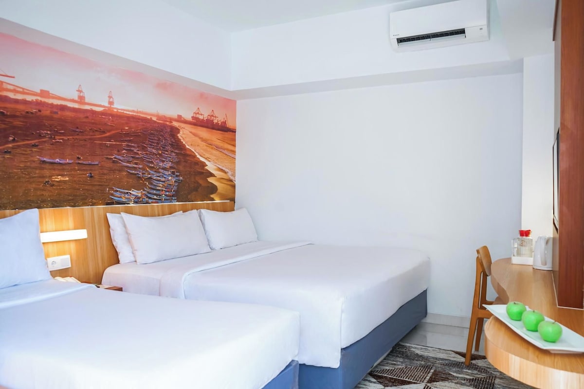 A hotel room with two beds, a desk with a chair, a small table with apples, and a wall-mounted air conditioner.
