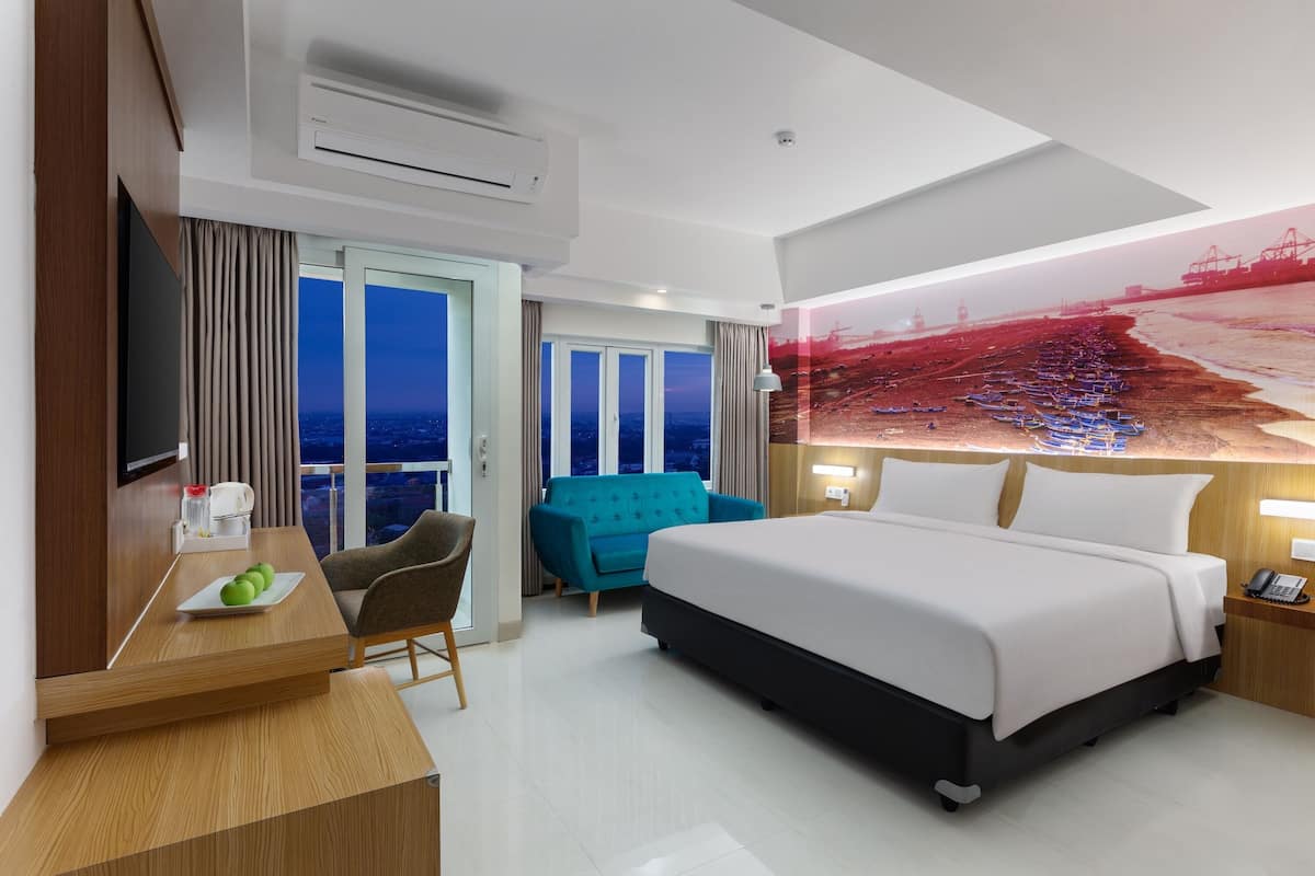 A modern hotel room with a large bed, a desk with a chair, a sofa, and a large window with a city view.