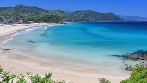 Ein Strand mit klarem türkisblauem Wasser, sandigem Ufer und grünen Hügeln im Hintergrund.