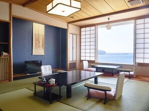 A traditional Japanese room with tatami flooring, a low table, seating, a television, and a view of the sea.