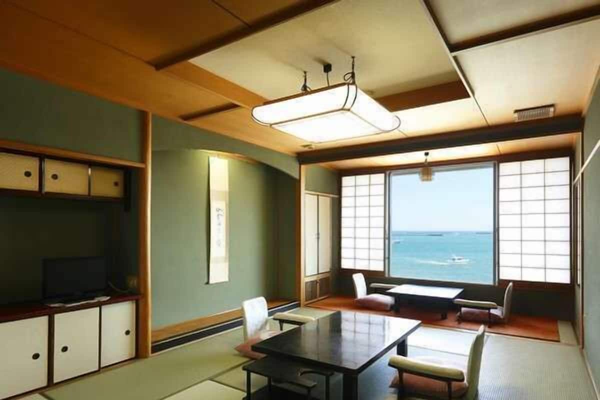 Une pièce de style japonais traditionnel, avec un sol recouvert de tatamis, une table basse, des chaises, un téléviseur et de grandes fenêtres donnant sur la mer.