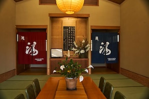 Een traditionele Japanse kamer met tatami vloerbedekking, een houten tafel en stoelen. Er zijn twee deuren met bordjes waarop 'Dames' en 'Heren' toiletten staan. Op de tafel staat een vaas met witte bloemen, en aan de muur hangt een ingelijst certificaat.