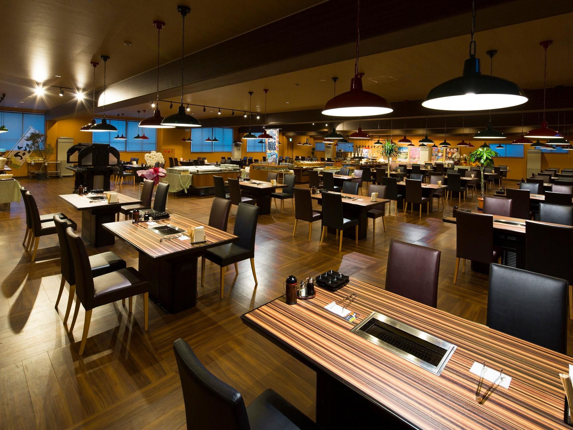 A spacious restaurant with multiple dining tables and chairs, overhead lighting, and a central bar area.