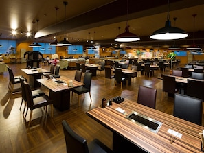 Een ruim restaurant met meerdere eettafels en stoelen, verlichtingsarmaturen aan het plafond en een bar in het midden.
