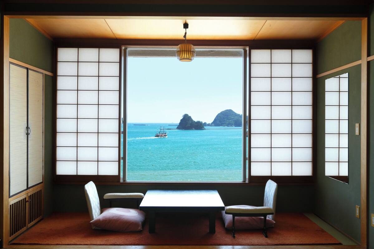 Une pièce de style traditionnel avec un sol recouvert de tatamis, une petite table basse, deux coussins et une vue sur la mer à travers des panneaux coulissants.