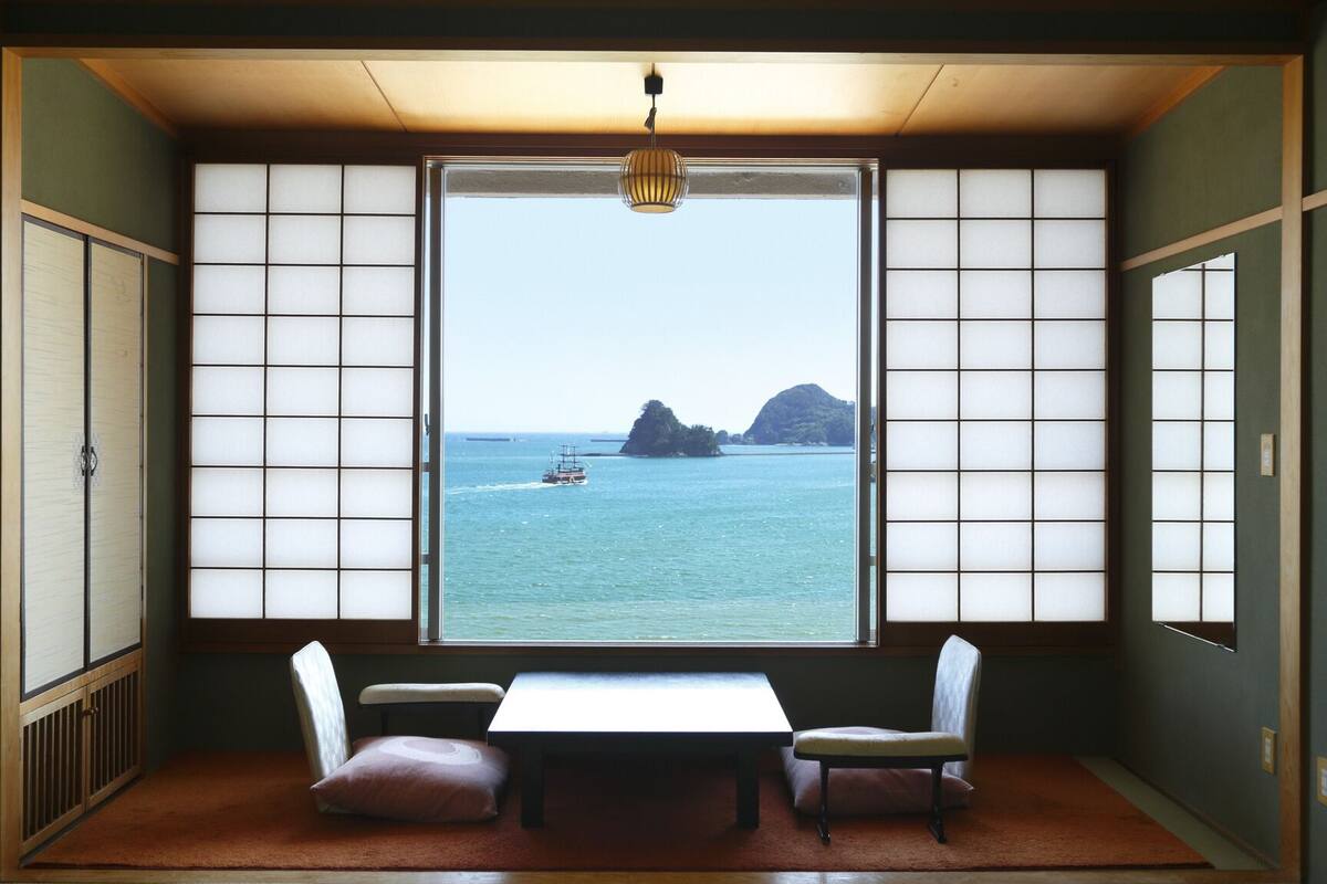 Une pièce dotée d’un sol recouvert de tatamis, d’une table basse, de deux chaises et d’une grande fenêtre avec des panneaux coulissants de type shoji.