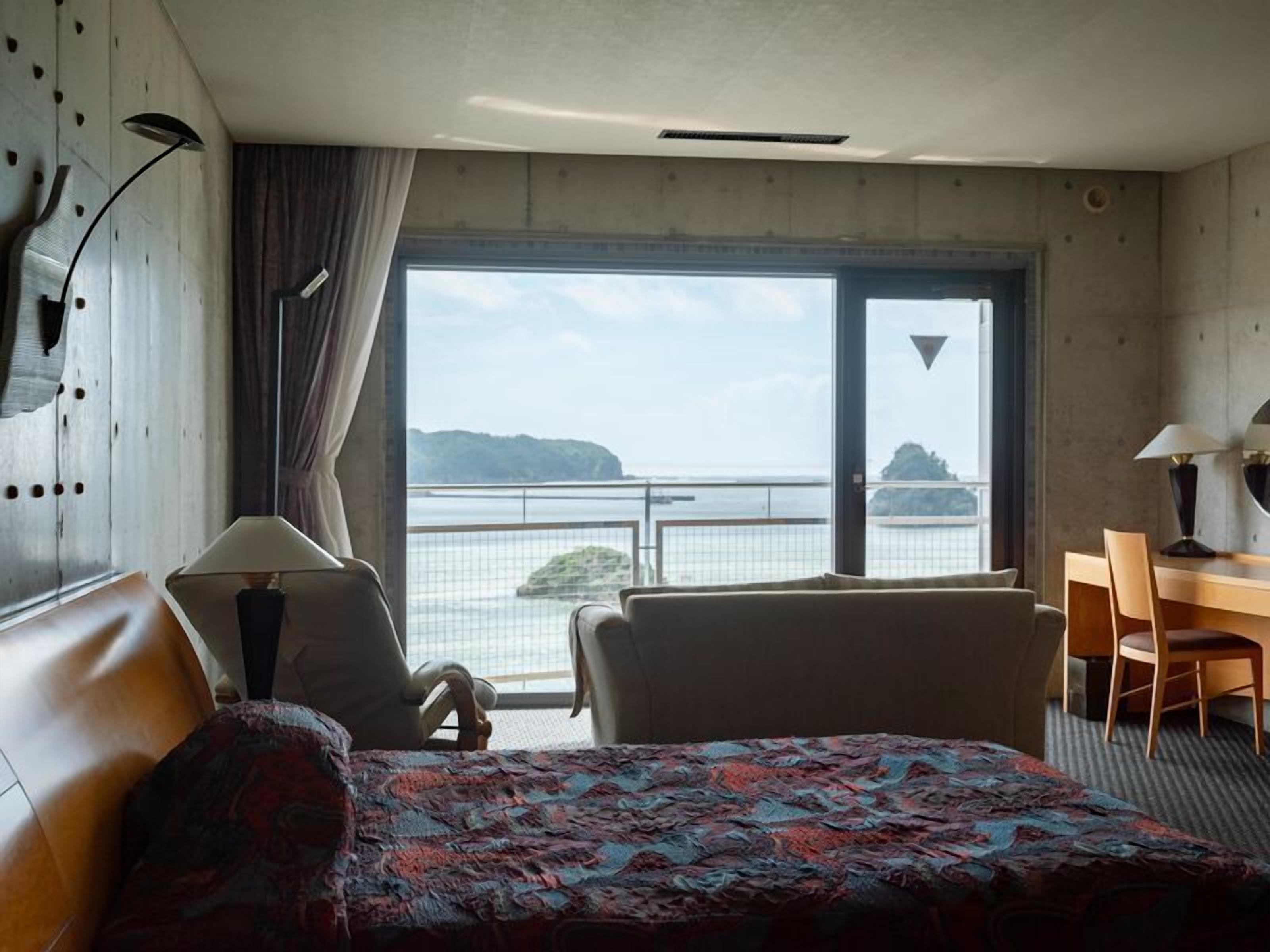 A hotel room with a bed, a desk, a chair, a lamp, and a view of the sea.